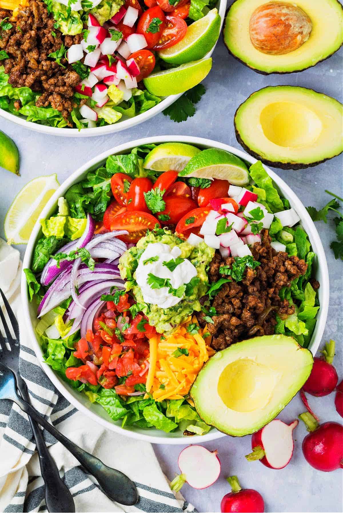 a bowl of keto taco salad with lettuce, tomatoes, taco meat, guacamole, radishes, cheddar, and Pico De Gallo.
