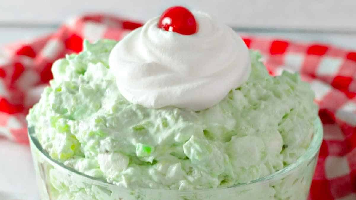 A bowl of green dessert with whipped cream and a cherry on top.