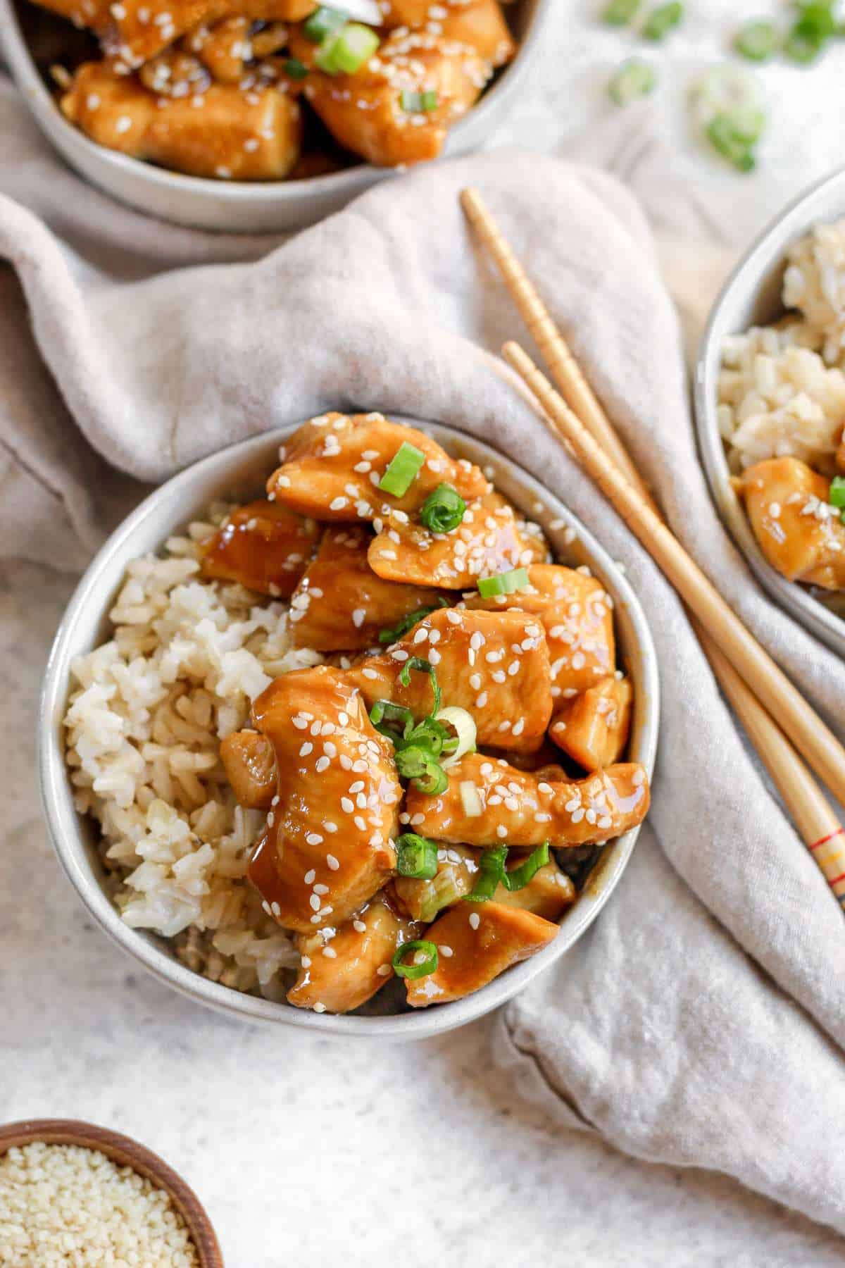 Gluten free chicken teriyaki sitting on top on rice in a round bowl with a tan tea towel and chopsticks on the side of the bowl. 
