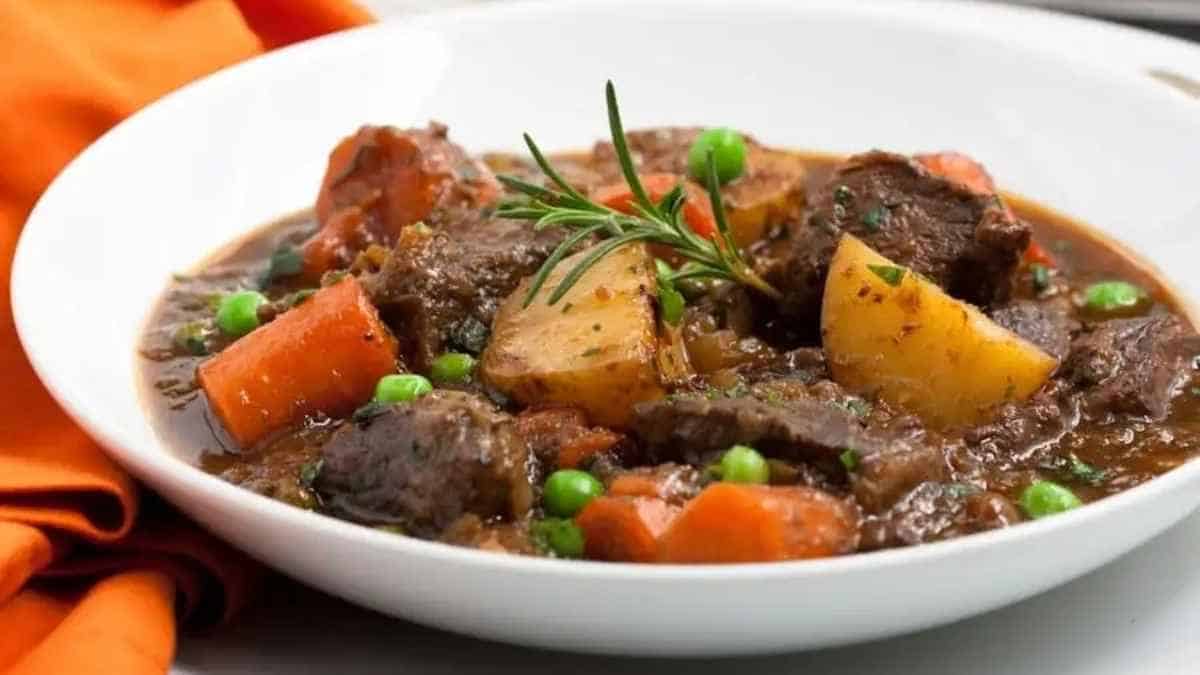 A bowl of beef stew with carrots, potatoes and peas.