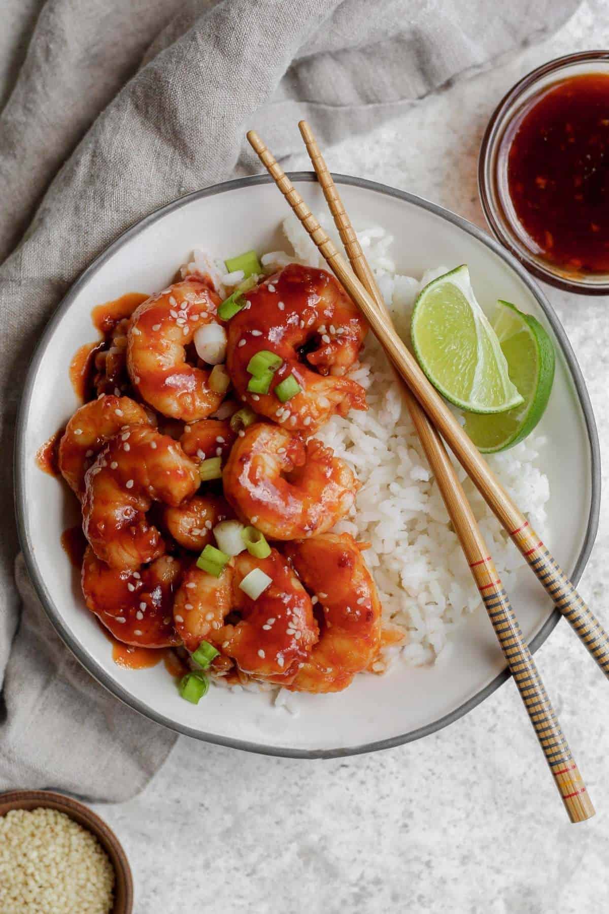 A bowl filled with white rice and topped with honey sriracha shrimp with a garnish of lime and chopsticks sitting on the side of the bowl. 
