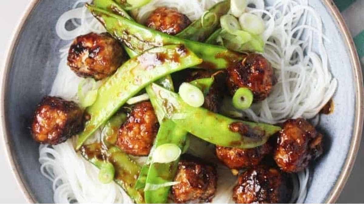 A bowl of noodles with meatballs and green peas.