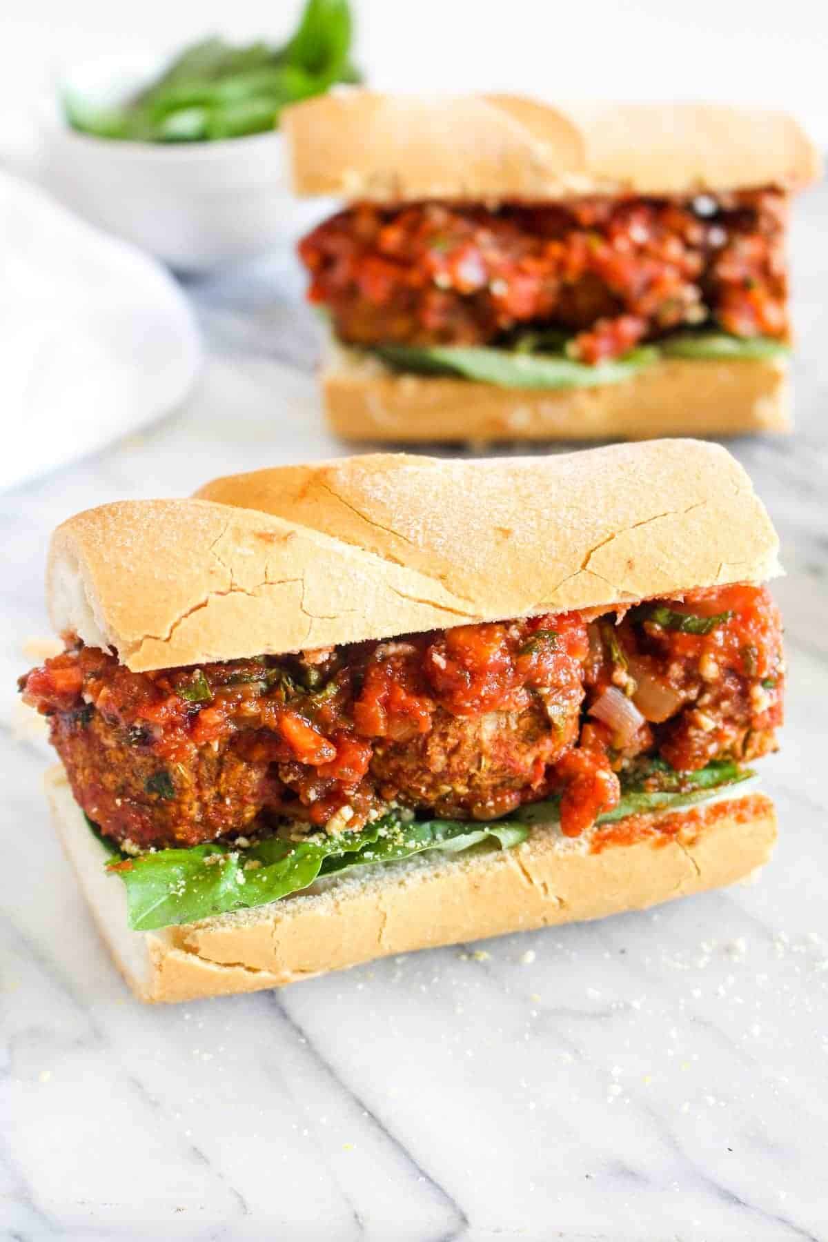 Two vegan meatball sandwiches with marinara sauce and basil on a white roll. 
