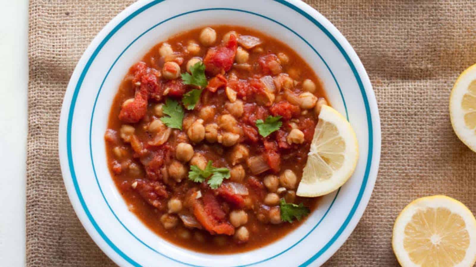A bowl of tomato and chickpea stew with lemon wedges.
