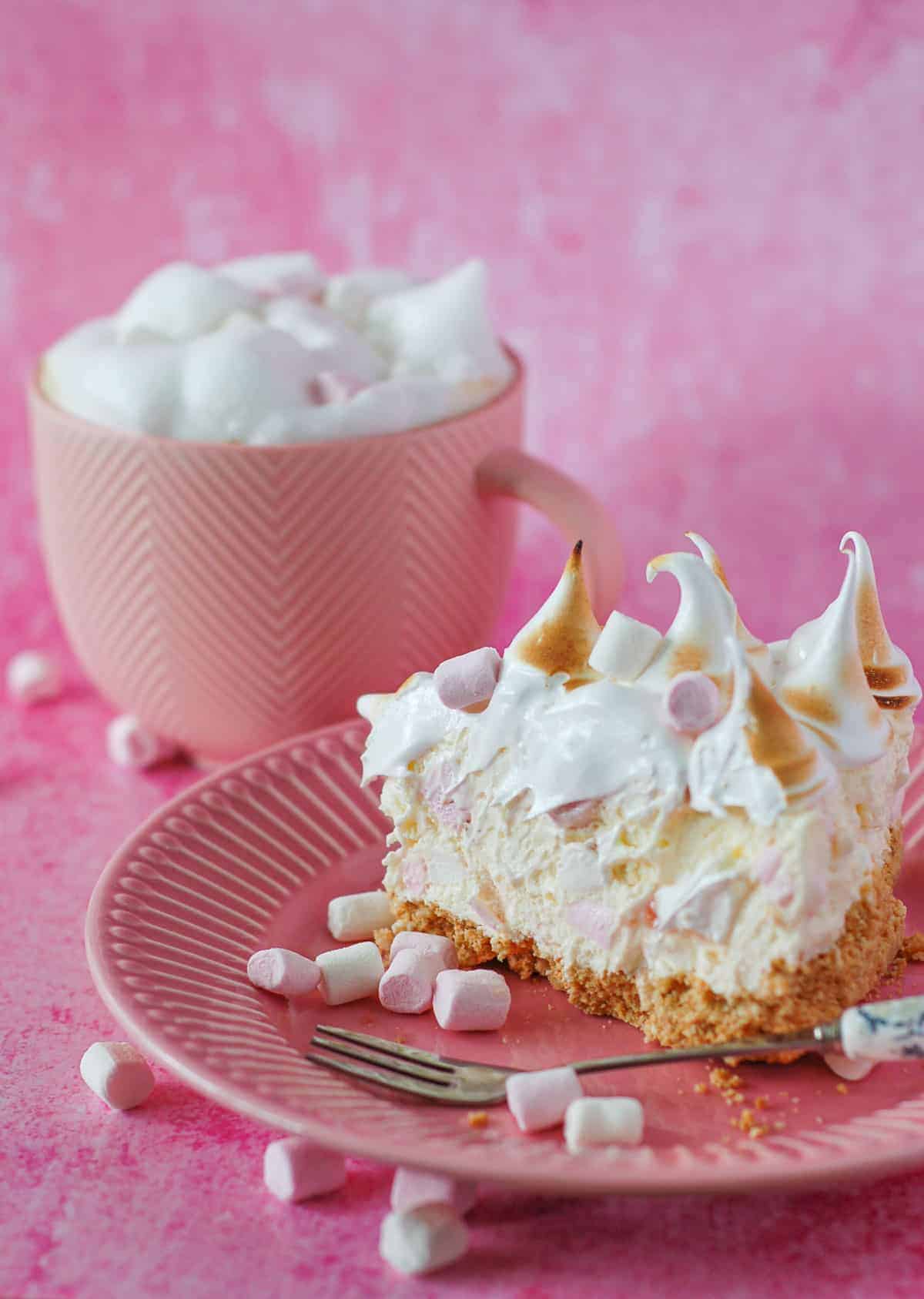 A slice of marshmallow tart on a pink plate with a mug of coffee in the background.
