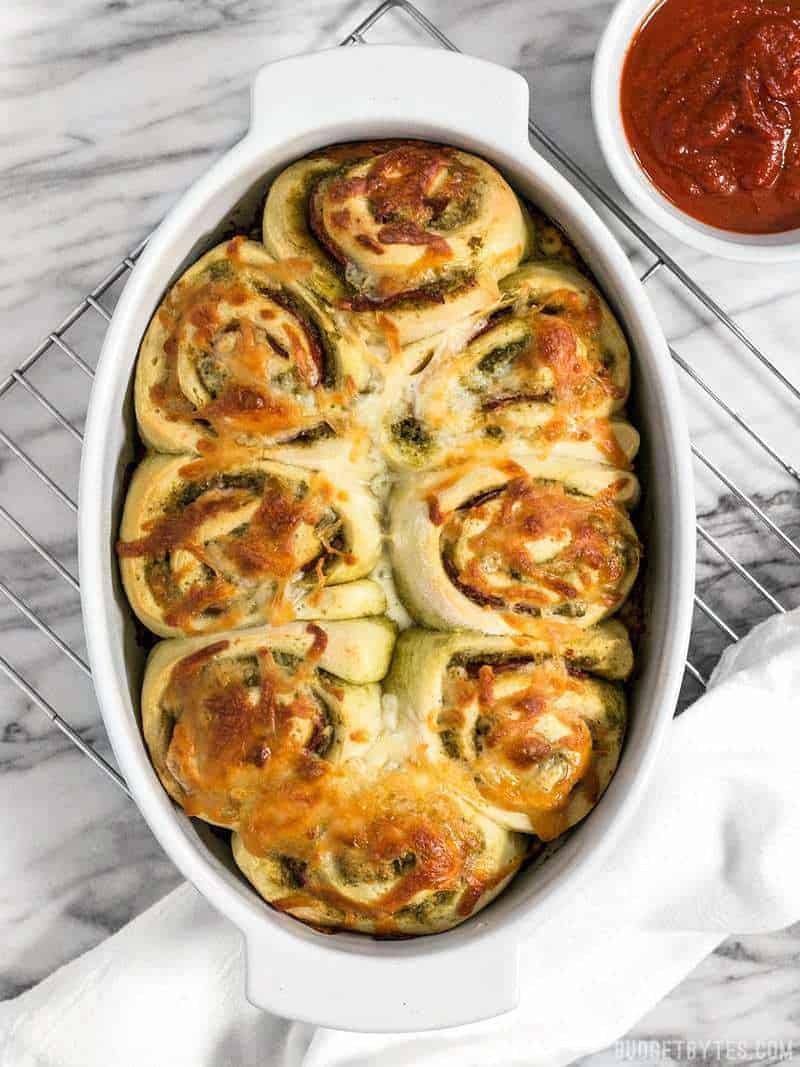 A baking dish full of Pesto Pizza Rolls on a cooling rack with pizza sauce on the side / These Pesto Pizza Rolls are a fun way to change up your pizza routine with swirls of cheese, pesto, and pepperoni, plus lots of crispy edges! 
