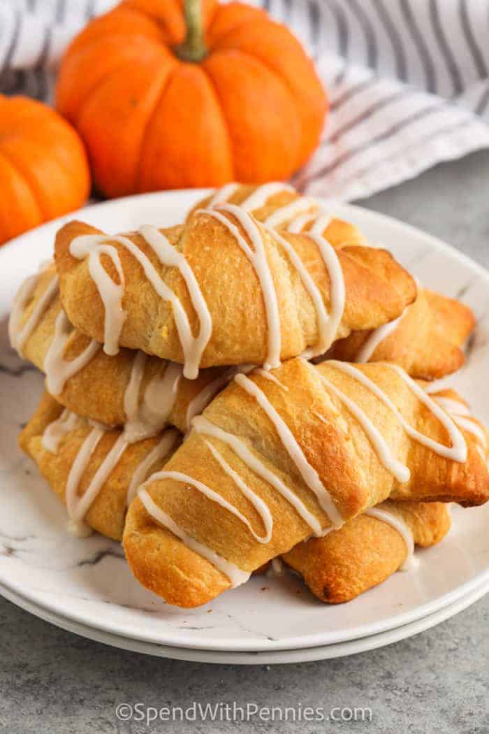 plated Pumpkin Pie Crescents with pumpkins in the back / A warm pumpkin pie crescents dessert is just what the family wants for dessert tonight! In just 20 minutes with pre-made crescent dough, these mini pumpkin pie crescents are ready to be devoured. Kids and adults alike all love these warm and flaky crescents with pumpkin puree and white frosting! Why not make a batch and enjoy them all week long as an after-school snack or late-night dessert? 
