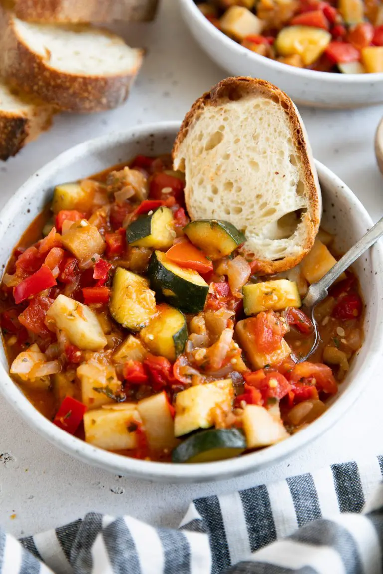 White shallow bowl filled with homemade Italian ratatouille and served with fresh toasted bread. 
