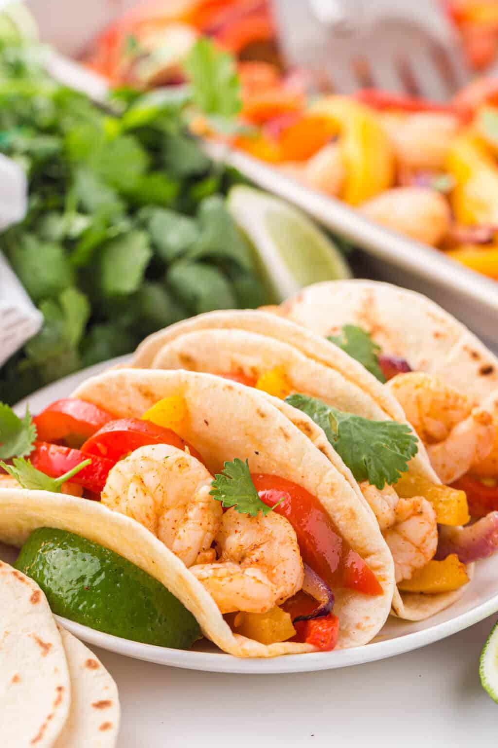 Flour tortillas filled with a roasted shrimp fajita recipe, with cilantro and a sheet pan in the background. 
