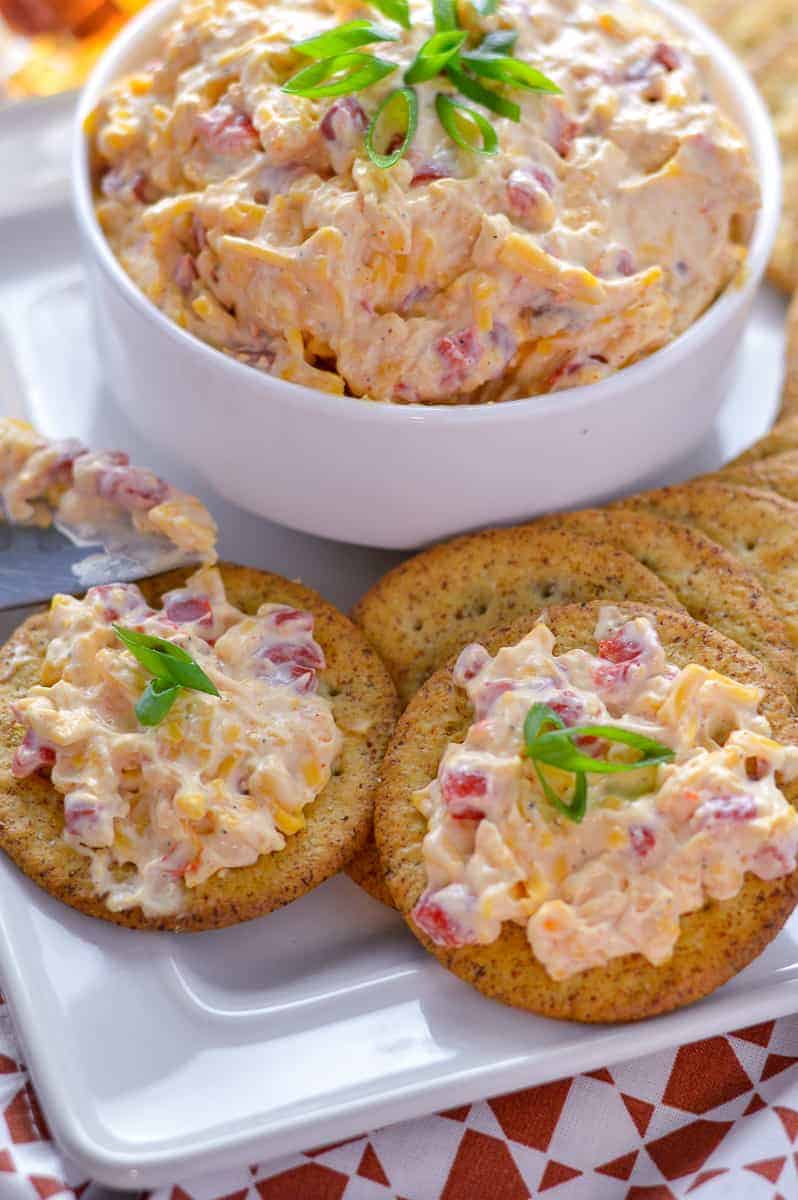 Close up view of pimento cheese spread on crackers with chopped green onion atop and knife with some spread on it.
