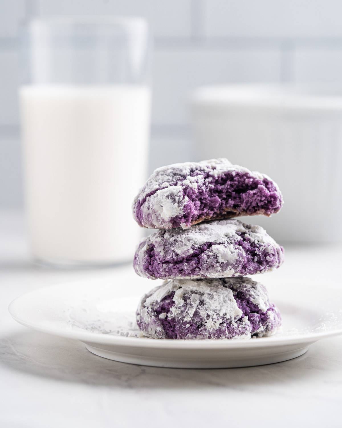 A stack of unique purple powdered sugar cookies on a plate next to a glass of milk.