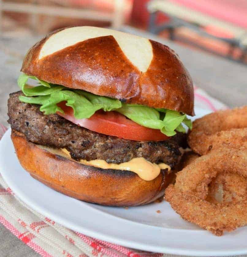 Grilled burger on a pretzel bun with a tomato, green lettuce, pink sauce on a white plate and onion rings peeking in the right side.
