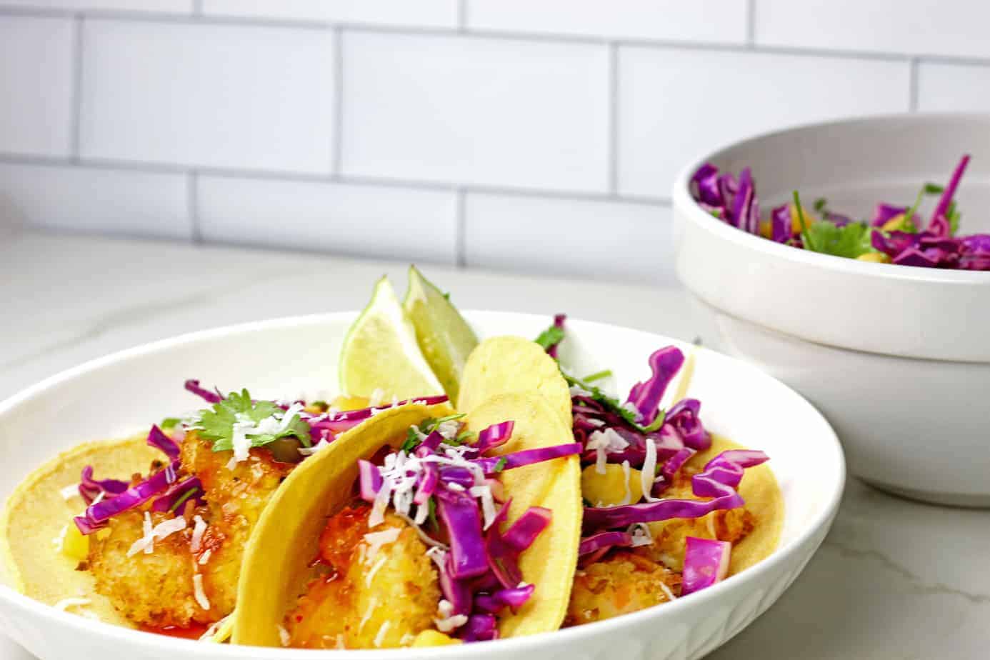 Three air fryer coconut shrimp tacos with a bowl of slaw in the background. 
