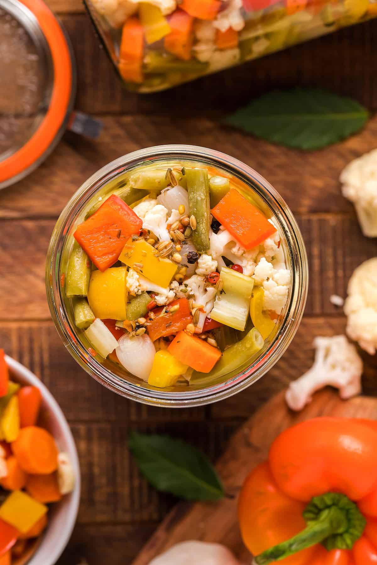 An overhead view of a jar of the picked vegetable relish on the top of a wood table. There is cauliflower and orange bell peppers on the table, too. 
