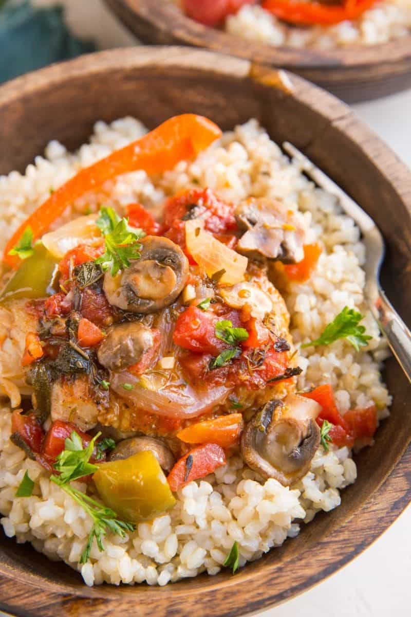 Wooden bowl with brown rice and chicken cacciatore with green bell peppers, tomatoes and onions and fresh parsley on top.
