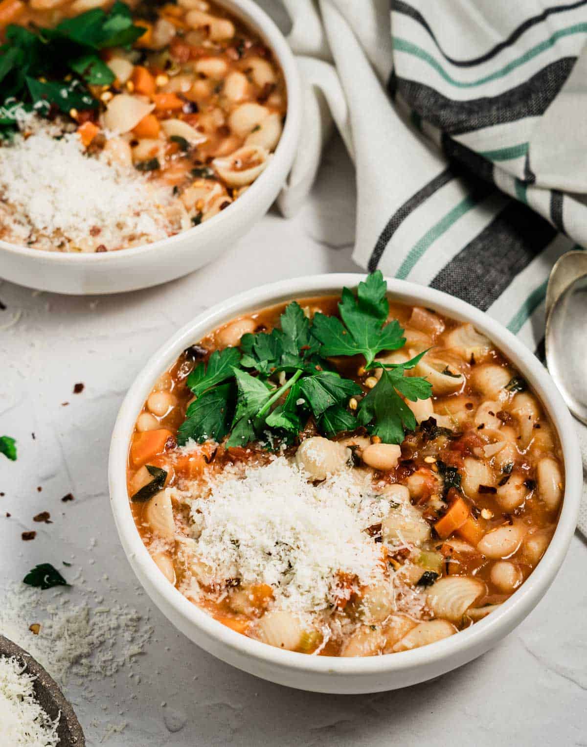 White bowl filled with gluten free pasta fagioli topped with grated parmesan and chopped parsley with a spoon on the side, a bowl of soup in the background and a striped napkin on the side. 

