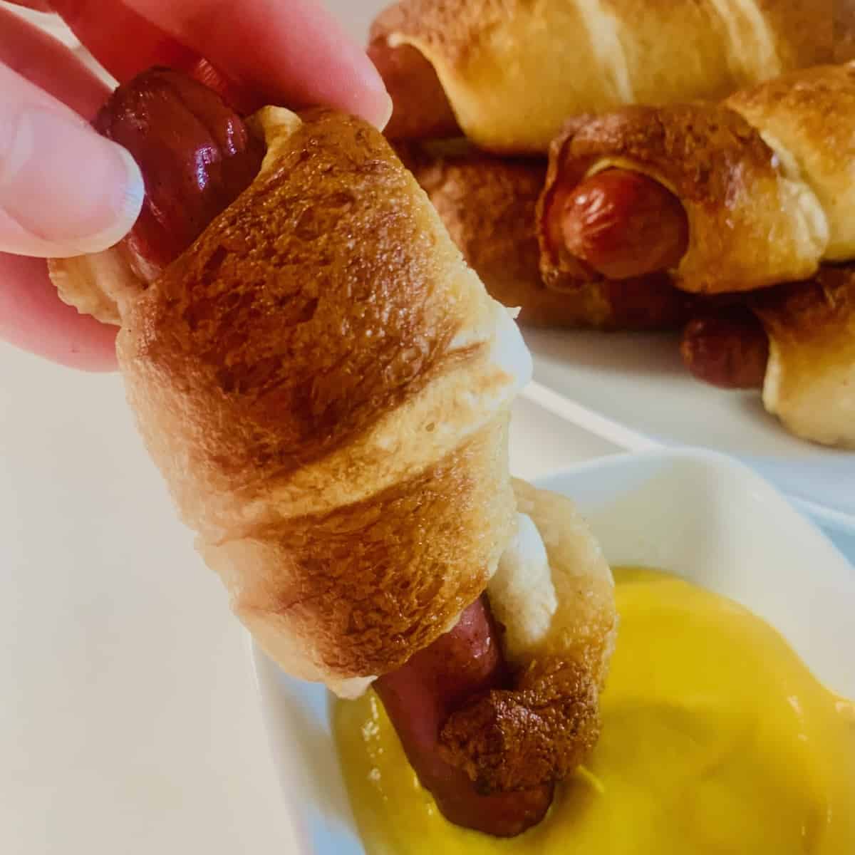 A gluten-free pretzel dog being dipped in mustard, with more gluten-free pretzel hotdogs in the background. 
