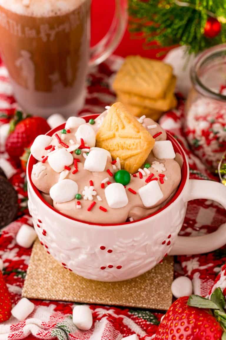 Close up photo of a white holiday mug filled with hot chocolate dip with marshmallows and sprinkles on top. A shortbread cookie is stuck in the top of the dip. 
