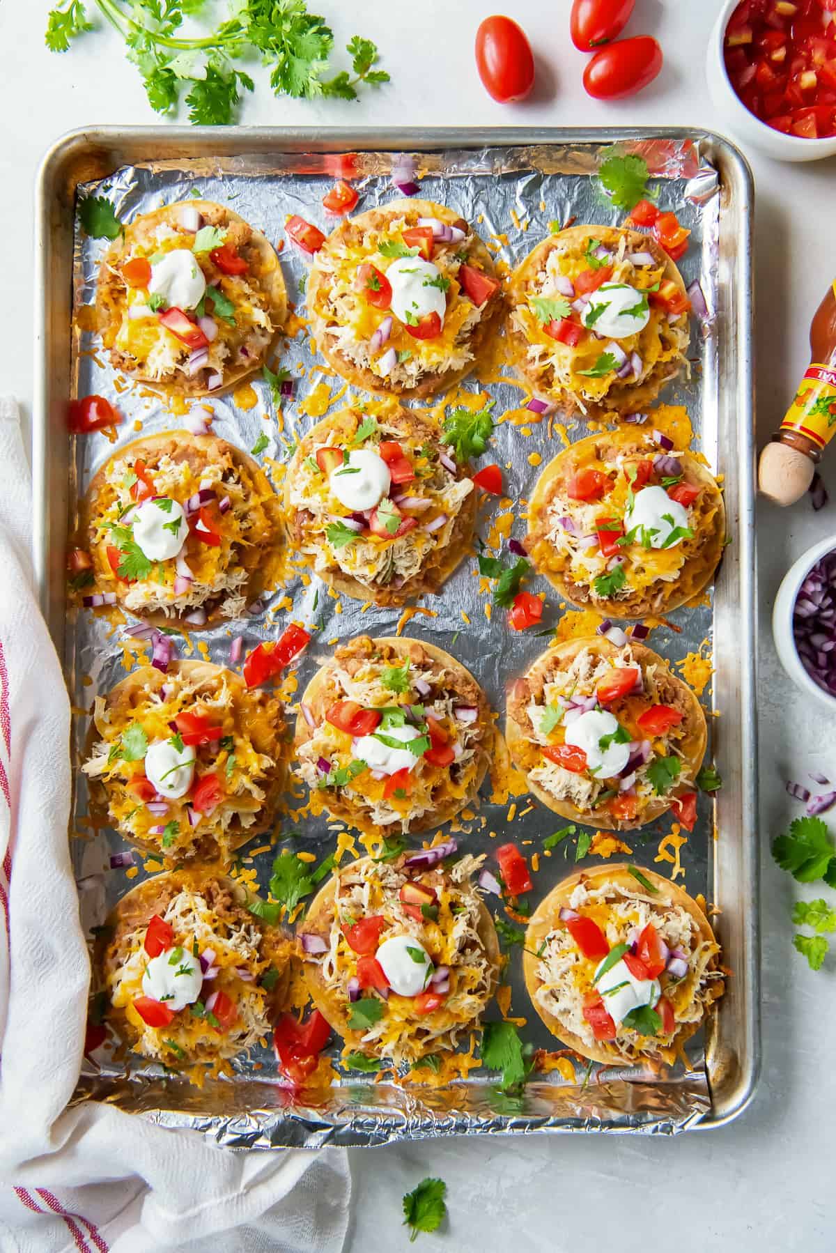 A top down shot of little tostadas with cheese, sour cream and tomatoes on a foil lined baking sheet. 
