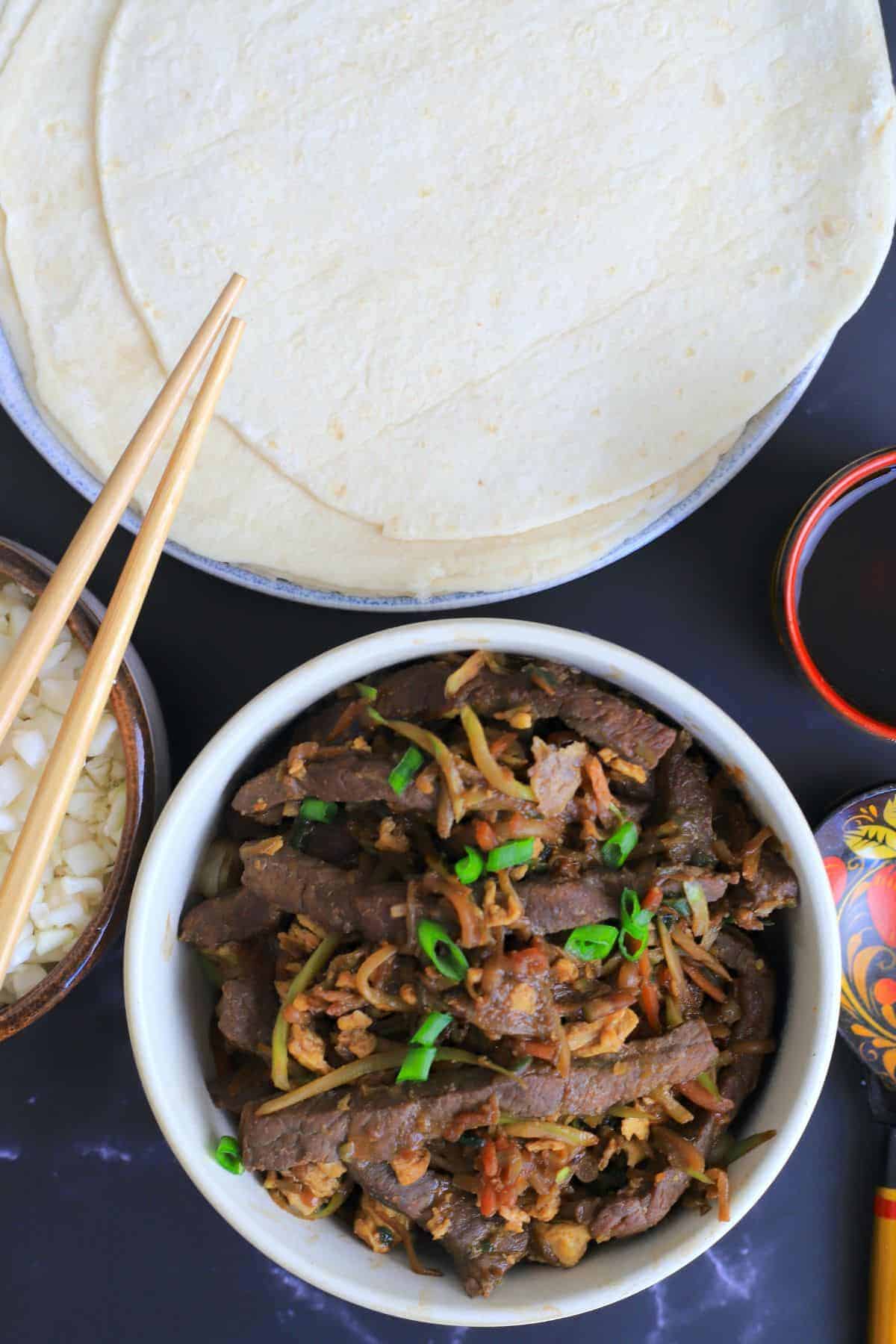 moo shu beef in a bowl next to a bowl with cauliflower rice and chopsticks with another dish with tortillas. 

