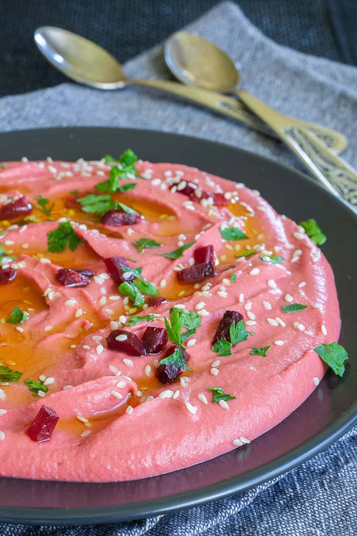 Pink hummus on a black plate, sprinkled with freshly chopped parsley, sesame seeds and small roasted beet pieces.
