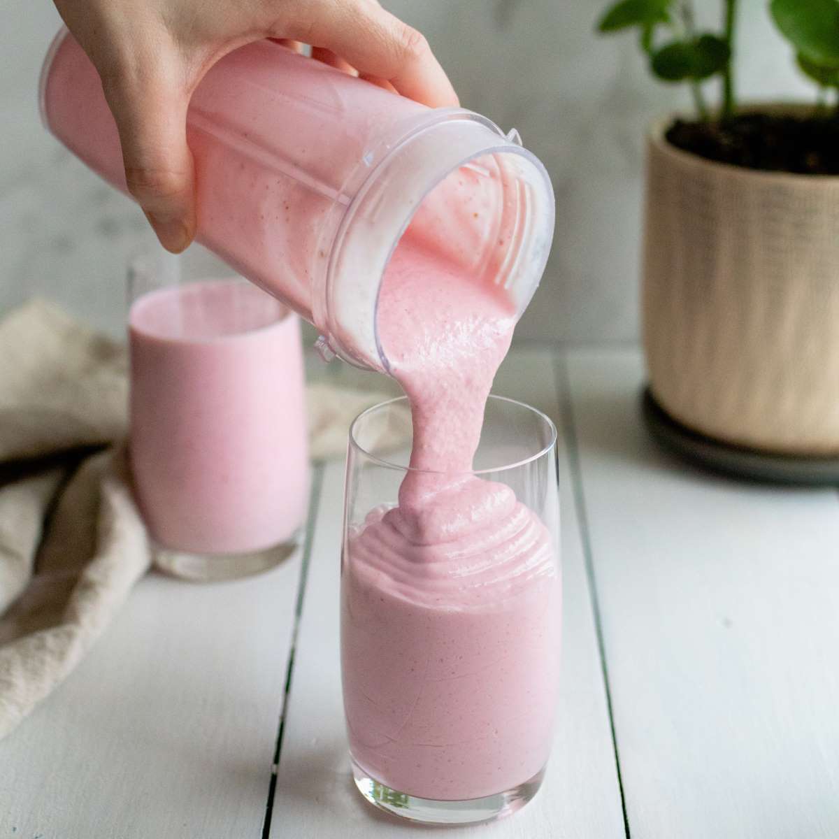 Pouring Strawberry Cottage Cheese Smoothie from a blender cup into a tall glass sitting on a wooden board. 
