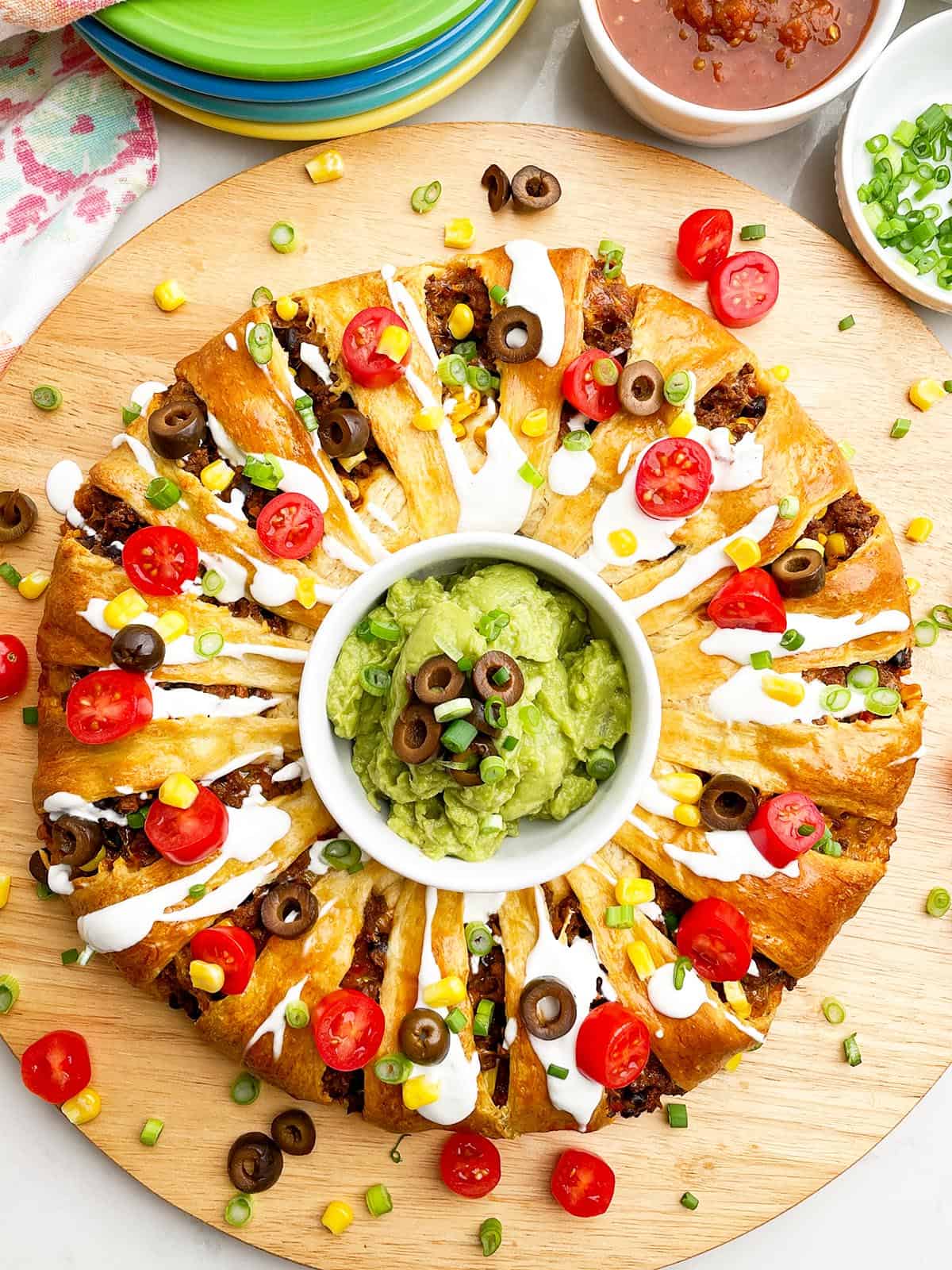 taco ring covered in toppings with guacamole in the center on a wooden cutting board.

