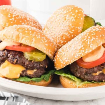 Two cheeseburgers with lettuce, tomatoes, pickles, and sesame seed buns on a plate.