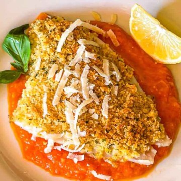 A breaded and baked fish fillet served on a bed of tomato sauce, garnished with grated cheese, a basil leaf, and a wedge of lemon.