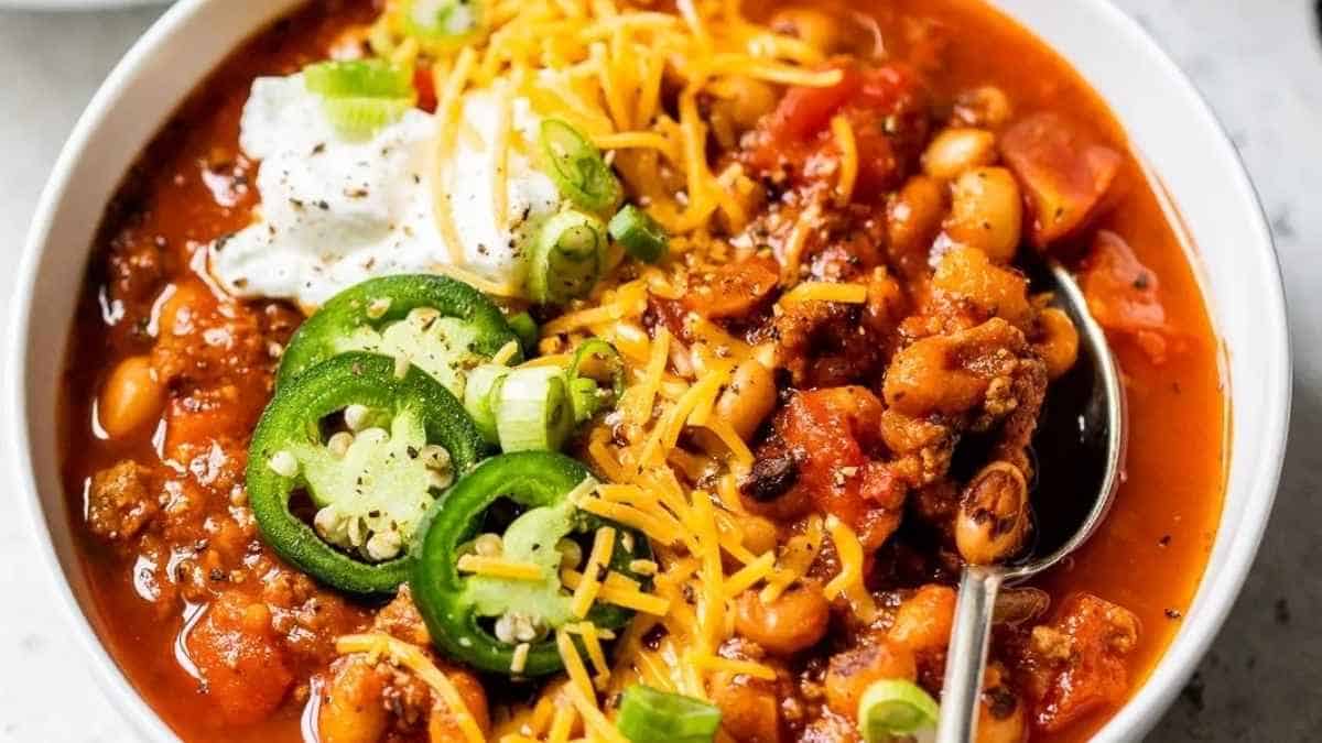 A bowl of chili topped with shredded cheese, sour cream, sliced jalapeños, and green onions.