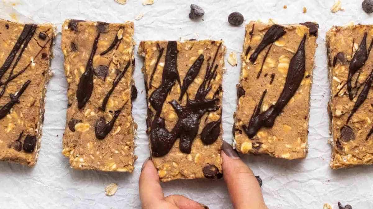 Three homemade granola bars with chocolate drizzle on a white surface, with a hand picking one up.
