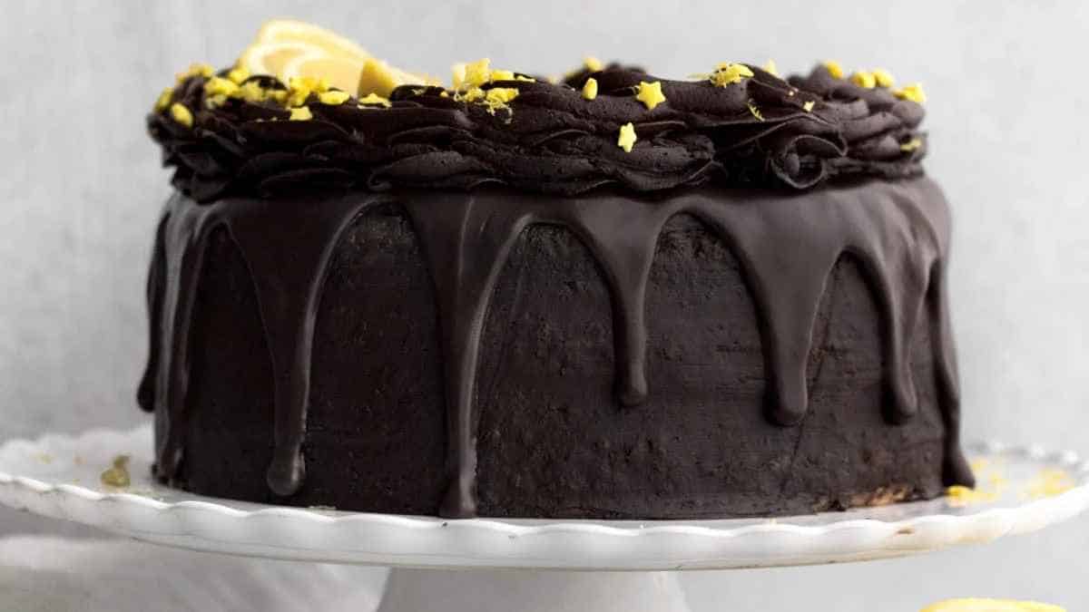 Chocolate cake with dark chocolate frosting, drizzled with ganache, and topped with lemon zest on a white cake stand.