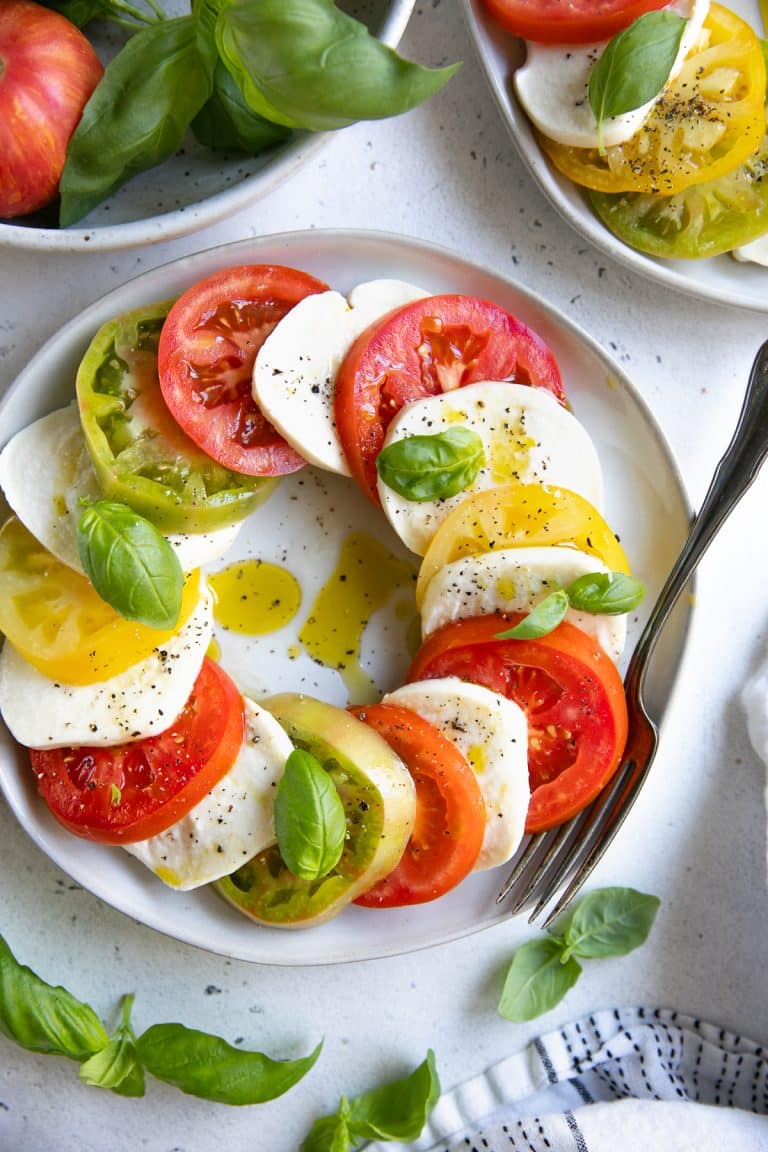 White Salad plate filled a caprese salad made of overlapping slices of fresh tomatoes and mozzarella cheese drizzled with olive oil and garnished with fresh basil. 
