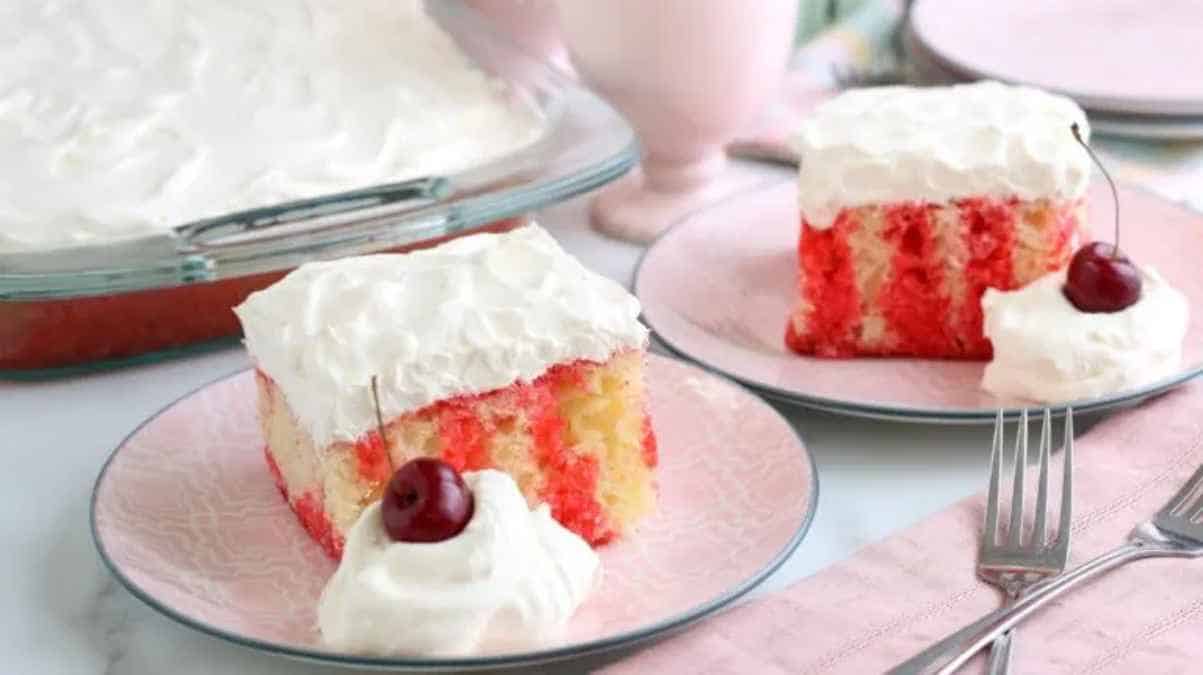 A slice of red and white poke cake with whipped topping on a plate, accompanied by a whole cake in a glass dish.