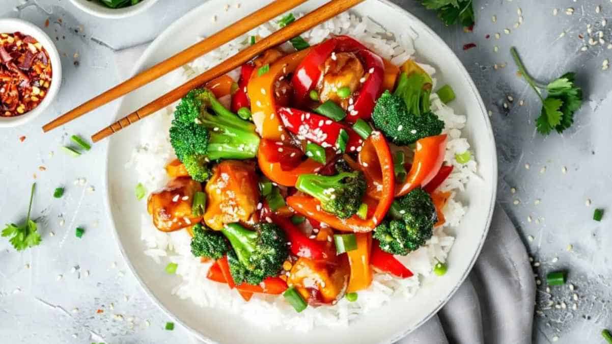 Colorful stir-fried vegetables and chicken served over white rice in a bowl with chopsticks.