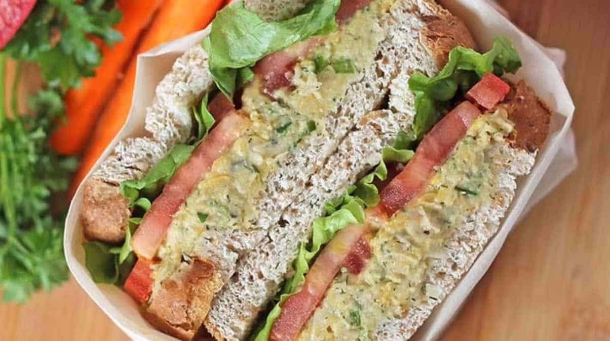 A chicken salad sandwich with lettuce and bacon on whole grain bread, served with carrots on the side.