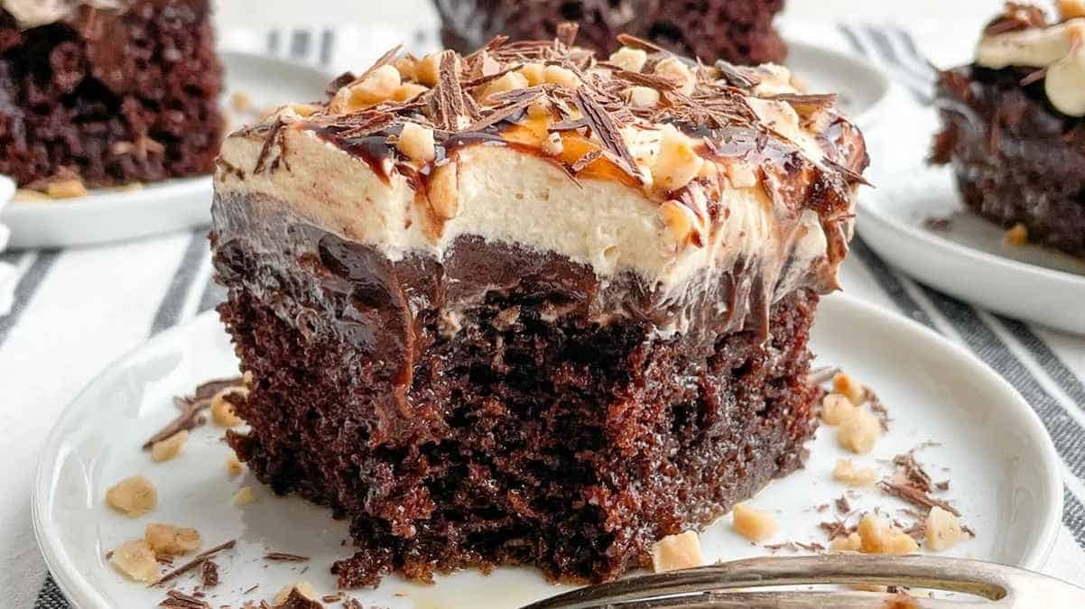 A slice of chocolate cake topped with creamy frosting, drizzled with chocolate syrup and sprinkled with nuts, served on a white plate.