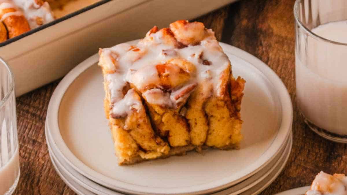 A piece of glazed cinnamon roll cake on a plate with a glass of milk in the background.