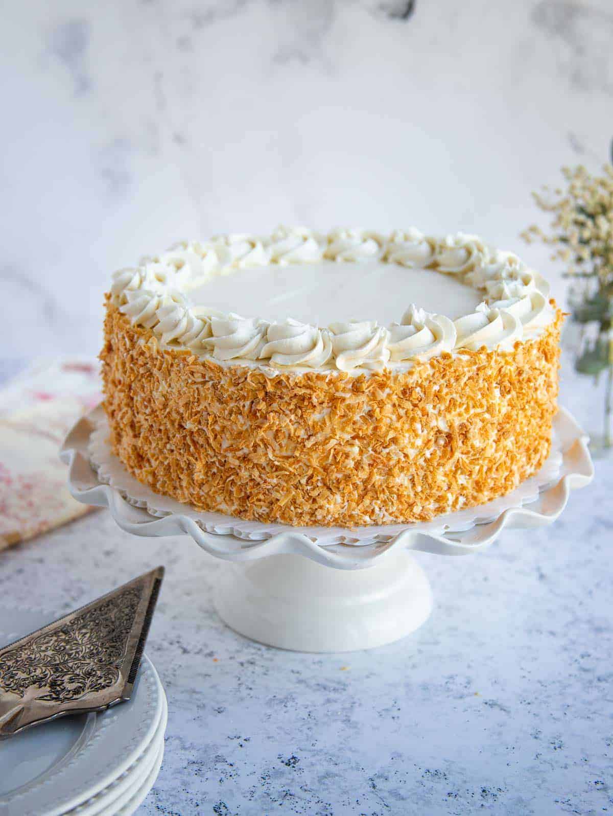 Coconut Layer Cake with Toasted Coconut Frosting on cake stand. Cake server and cloth napkins on the left of the cake stand. 
