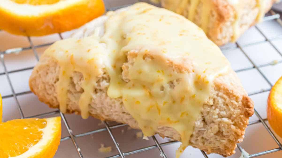 Freshly baked orange scone with glaze on a cooling rack, surrounded by orange slices.