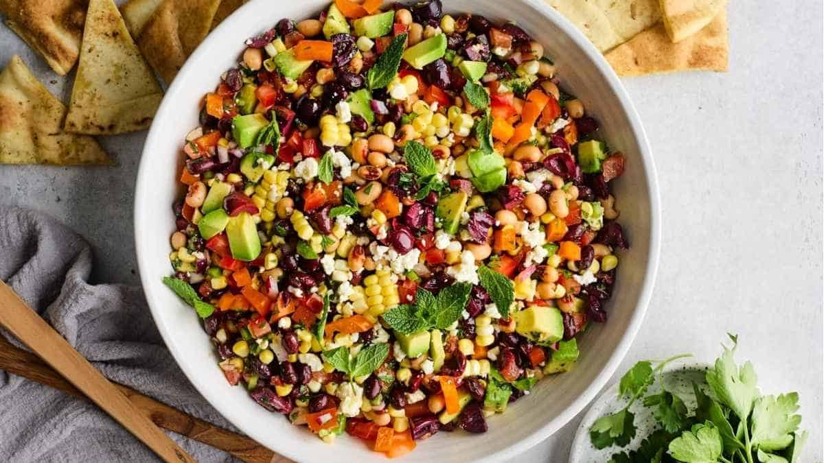 A colorful bowl of mixed bean salad with avocado, herbs, and pita chips on the side.