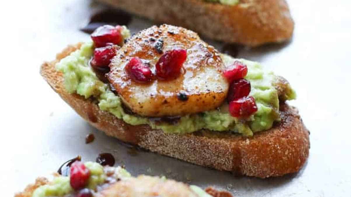 A slice of toast topped with avocado, a seared scallop, and pomegranate seeds.