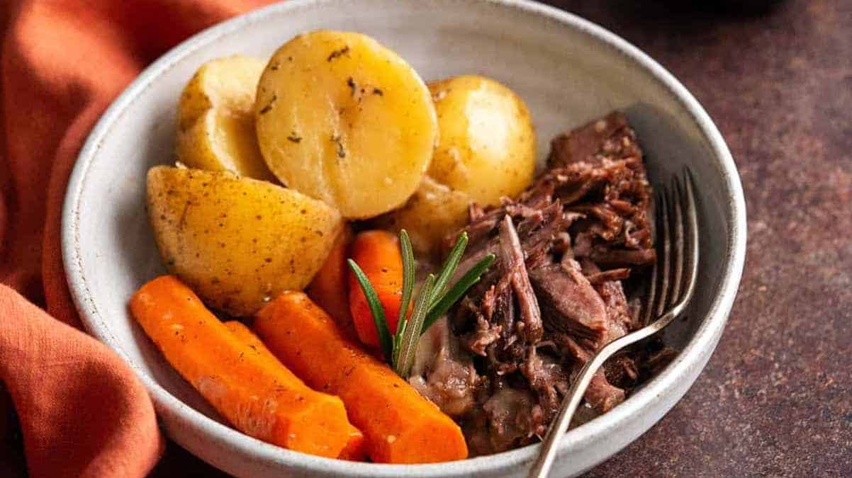 A plate containing a pot roast with carrots, potatoes, and a sprig of rosemary.