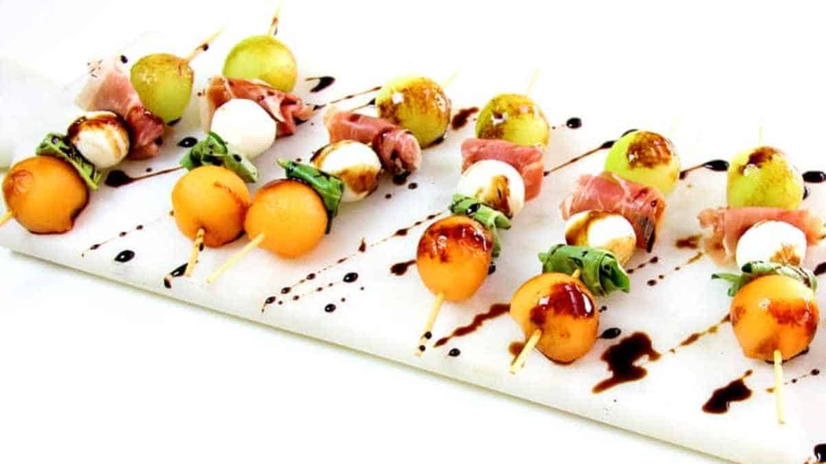 Assorted melon and prosciutto appetizers with balsamic glaze on a white surface.