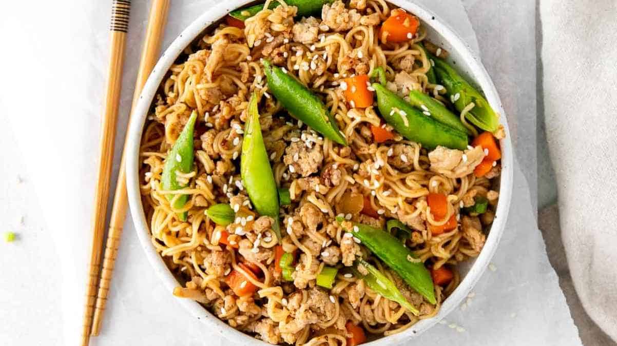 Bowl of stir-fried noodles with vegetables and ground meat accompanied by chopsticks.