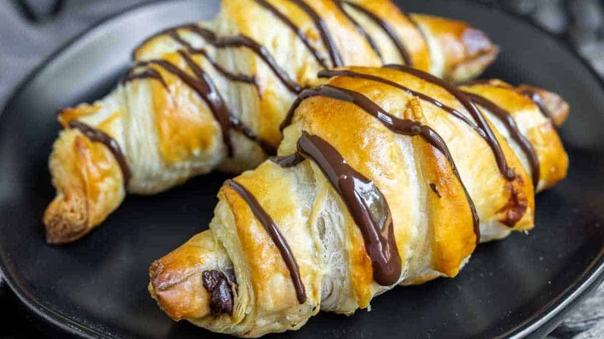 Freshly baked croissants drizzled with chocolate sauce on a black plate.