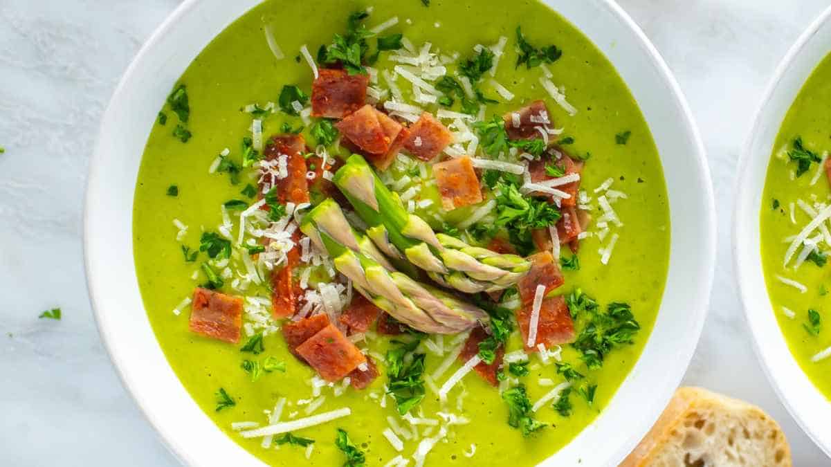 A bowl of creamy asparagus soup garnished with diced ham, shredded cheese, and asparagus tips, served with a side of bread.