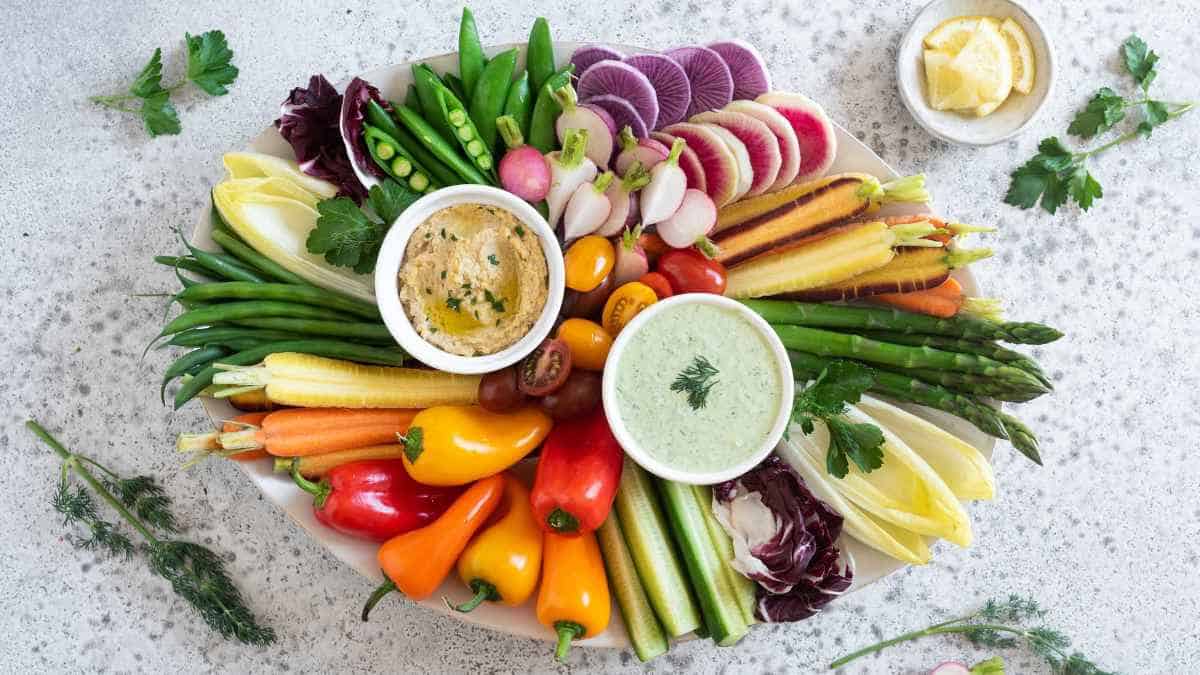 An assortment of fresh vegetables neatly arranged around two bowls of dip on a white surface.