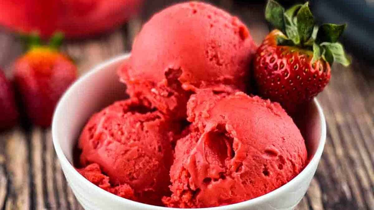 Three scoops of strawberry ice cream in a white bowl with a fresh strawberry on the side.