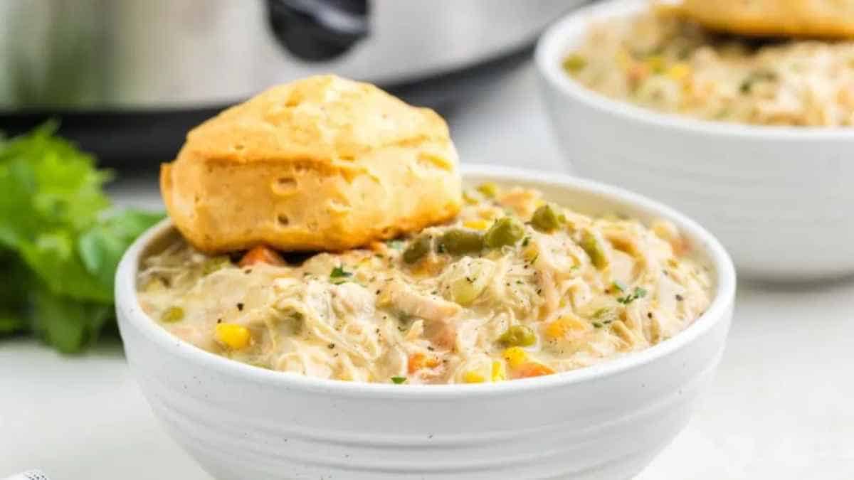 A bowl of creamy chicken noodle soup with vegetables, garnished with parsley, and accompanied by a biscuit.